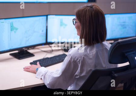 Side-rear view of a woman with short brown hair and glasses working at a desk with three computer monitors displaying maps and data systems inside a s Stock Photo