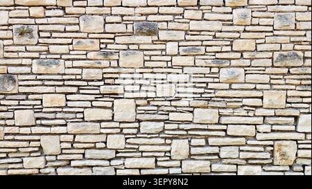 Close-up of a beige and gray natural stone wall featuring various sized rectangular and square stones arranged in an irregular pattern with visible mo Stock Photo