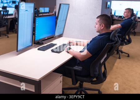 Side profile of a male security professional sitting at a desk and using a keyboard and mouse with three computer monitors displaying data and system Stock Photo