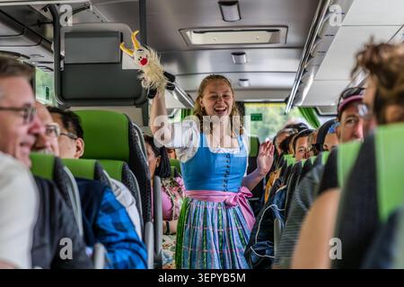 Tour guide Stephanie Hinterbuchner in a dirndl laughs and shows the passengers Goatherd, a soft toy goat on a packed coach; several passengers laugh and sing along with her. Photograph taken on Wolfgangseestraße in the Salzkammergut, Salzburg, Austria Stock Photo