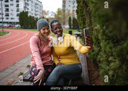 Diverse friends taking selfie sitting on bench outdoors Stock Photo