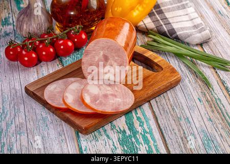 Sliced smoked sausage on wooden board with cherry tomatoes, garlic, green onions, yellow pepper, and oil on rustic table. Ready for sandwich or snack Stock Photo