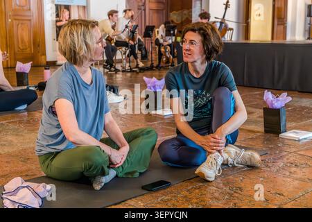 Andrea Stockhammer (right) and Angela Berg (left) are taking part in the ‘Pop Up Yoga – Mozart Edition’ event at the DomQuartier in Salzburg. The director is sitting on a yoga mat talking to the journalist; a chamber ensemble is playing live in the background. The event was part of the Mozart 270 programme to mark the 270th anniversary of the composer’s birth (1756–2026). Domplatz, Salzburg, Austria Stock Photo