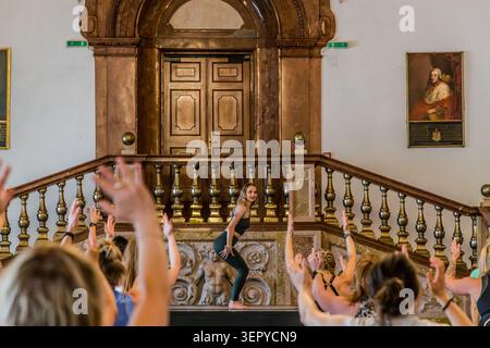 A yoga teacher is holding a balancing pose on stage in front of the grand Baroque staircase of the Archbishop’s Residence; yoga participants behind her are stretching out their hands. The gilded balustrades and stucco reliefs on the staircase date from the early 18th century. DomQuartier, Salzburg, Austria Stock Photo