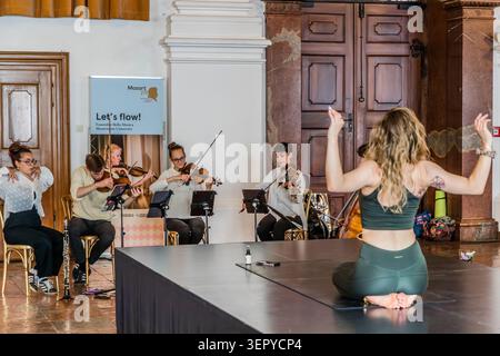 A yoga teacher demonstrates an arm movement on stage; behind her, the Bella Musica ensemble from the Mozarteum University in Salzburg is performing. The roll-up banner identifies the event as “Let’s flow! – Ensemble Bella Musica / Mozarteum University” as part of Mozart 270. DomQuartier, Salzburg, Austria Stock Photo