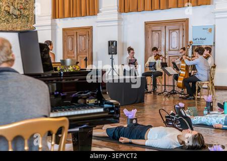 Overview: on the left, a pianist at a concert grand piano with singing bowls and an assistant; on the right, the Bella Musica ensemble from the Mozarteum University in Salzburg in front of the ‘Let’s flow!’ banner; Yoga participants are lying in Savasana on mats. Mozart 270 programme at the DomQuartier, Salzburg, Austria Stock Photo