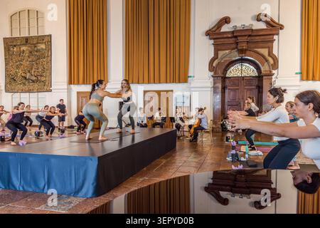 Two yoga teachers are demonstrating a balancing exercise together on the stage, holding hands; in the background, the string section of the Ensemble Bella Musica is playing, whilst participants in the hall follow along with the pose. Ballroom of the Archbishop’s Residence, DomQuartier, Salzburg, Austria Stock Photo