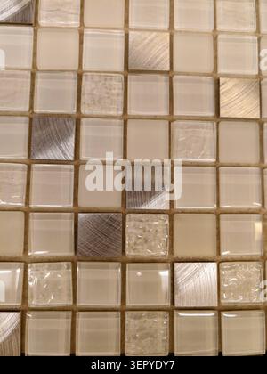 Close-up view of a mosaic tile surface composed of beige ceramic squares, textured metallic inserts and translucent glass pieces. The grid is set in l Stock Photo