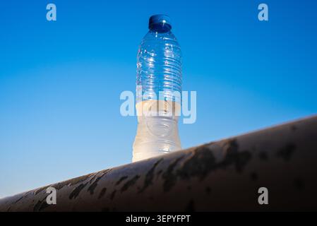 A clear plastic bottle stands upright on a dark rocky surface under bright sunlight. Stock Photo