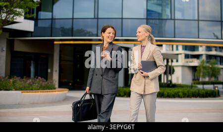 Two businesswomen chat and walk together outside a modern office building. They hold a briefcase and a tablet while dressed in suits. It is daytime an Stock Photo