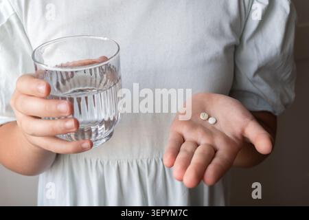 Girl in a dress take the two pills and glass with water in the hand. Healthcare from pharmacy. Kid sit at home because of illness. Stock Photo