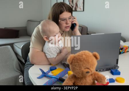 Young mother sits at a table holding her toddler on her lap while talking on a smartphone and working on a laptop. Soft natural light fills a living room Stock Photo