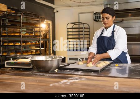 African American woman baker shaping dough rolls on bakery bench with mixing bowl, copy space Stock Photo