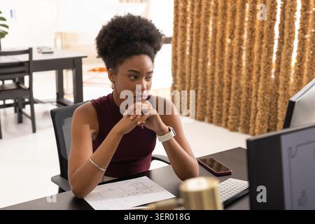 African American woman wearing sleeveless maroon top working at desk with floor plan and monitor Stock Photo