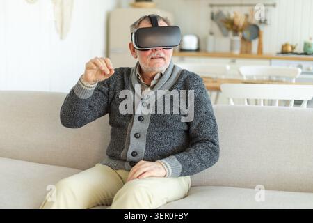 Excited mature senior man wearing using virtual reality metaverse VR glasses headset at home. Grandfather touching air during VR experience on virtual reality helmet. Simulation hi-tech videogame Stock Photo