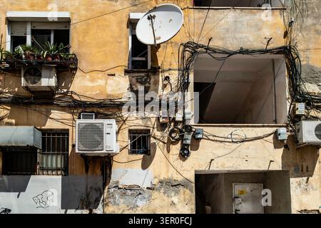 A tangled mess of electrical wiring, satellite dish, and air conditioners on a weathered yellow building facade in Tirana, Albania. Stock Photo