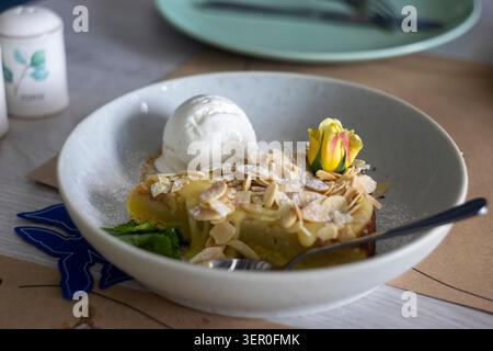 A slice of warm apple strudel or pie topped with almond flakes and powdered sugar, served in a ceramic bowl with a scoop of vanilla ice cream, garnish Stock Photo