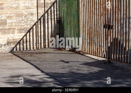 A weathered corrugated metal fence casts long dramatic shadows across the pavement beside a stone wall in bright sunlight, Avignon, France Stock Photo