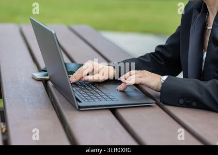 A person types on a laptop while seated at an outdoor table in a park. The grass is green and it is daytime. The setting looks casual and focused on w Stock Photo