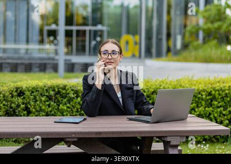 A woman is sitting at a wooden table outside in an office area. She talks on her phone while looking at her laptop and has a tablet beside her. It is Stock Photo