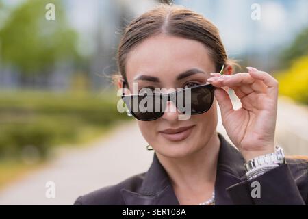 A woman adjusts her sunglasses and smiles while walking along a city path. She is dressed in a black suit and stands against a backdrop of green plant Stock Photo