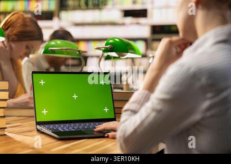 Isolated chroma key on laptop screen at library wooden table, students shown at desk learning in group study. Peer creating mockup space and screen replacement for digital learning. Stock Photo