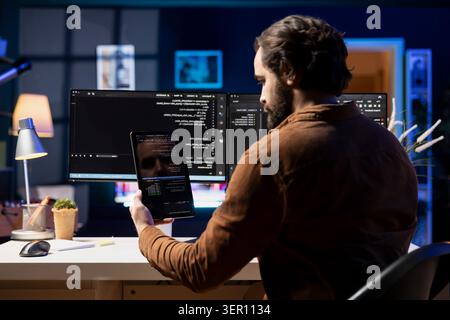 Programmer doing software debugging using tablet, checking glitches in lines of code displayed on computer monitors. Man working from home fixing coding issues using portable digital device. Stock Photo