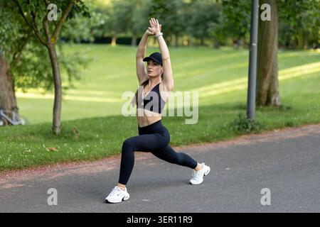 A woman is practicing yoga in a park. She stretches her arms and legs while wearing workout clothes. There are trees and grass around her. It is dayti Stock Photo