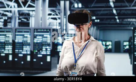 Older computer scientist in modern server hub wearing virtual reality headset, performing racks maintenance. Stock Photo