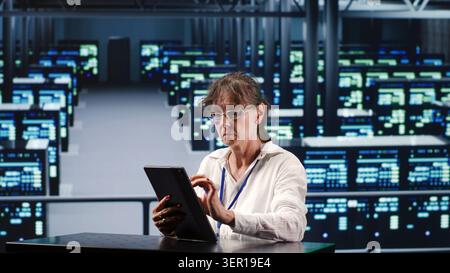 Competent manager in data center housing high-end devices capable of quickly and efficiently performing complex computations and data analysis, using tablet to fix issues. Stock Photo