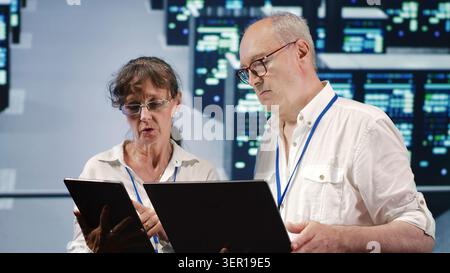 Data storage company executives looking around data center, using laptop and tablet to crosscheck contingency plan and assess high-tech workspace units in need of fixing, preventing liabilities. Stock Photo
