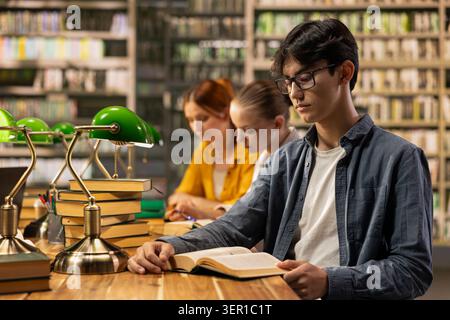 Young man with glasses reading open book at wooden library table, surrounded by green lamps and academic study materials. Asian student consulting textbooks resources near classmates at desk. Stock Photo