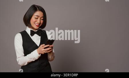Hospitality industry worker using smartphone app over grey background, looking at online information to confirm room reservations, asian receptionist browses websites. Stock Photo