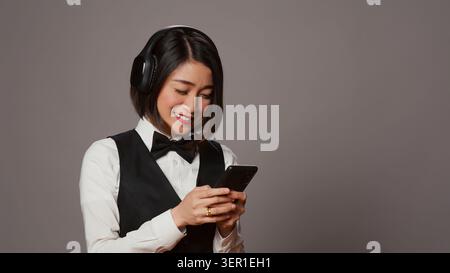 Front desk staff playing music on headphones using phone app, enjoying songs on radio and dancing in studio, asian employee having fun listening to modern tunes on headset. Stock Photo