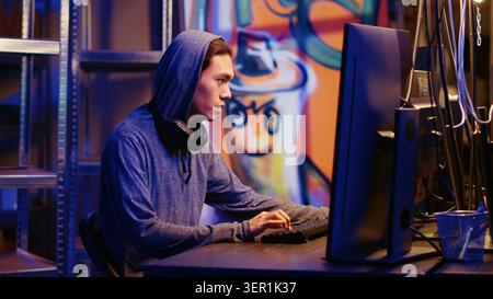 Happy Asian hacker in graffiti painted hideout having burst of joy after managing to steal valuable data, using phishing technique that tricks users into revealing sensitive information. Stock Photo
