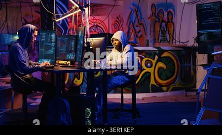 Team of hackers typing fast on keyboards in graffiti painted hideout, rushing to finish developing spyware software in order to use it to gather data from users computers without their knowledge. Stock Photo