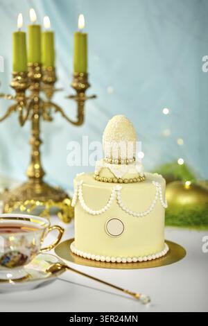 Small easter cake decorated with white chocolate egg, golden beads and white pearls, lit green candles and teacup in background, festive atmosphere Stock Photo