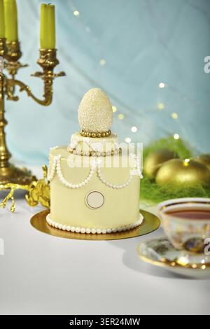 Small easter cake decorated with white chocolate egg, golden sprinkles, and white pearls on light blue festive background with golden candle holder an Stock Photo