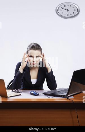 Office portrait of beautiful young business woman with headache sitting between tow laptops and massaging her temples Stock Photo