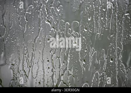 Close-up view of rain streaks and water droplets on a glass window, creating a moody gray texture and blurred background that evokes weather, reflecti Stock Photo