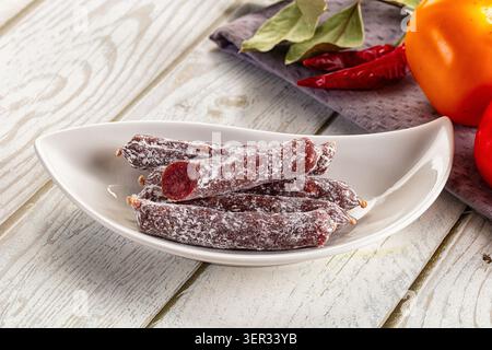 Traditional Spanish fuet sausage on white plate with peppers persimmon on rustic wooden table. Dried cured meat with white mold, red chili peppers, ba Stock Photo