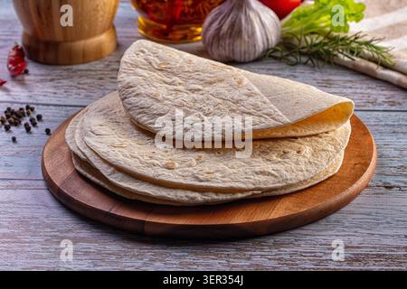 A stack of thin flatbreads, such as tortillas or lavash, on a round wooden board. In the background are fresh ingredients like garlic, herbs and veget Stock Photo