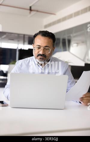 Focused Indian man comparing printed documents with digital data Stock Photo