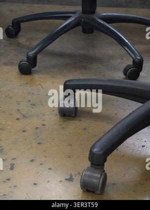 Close-up view of multiple black office chair bases and rolling casters resting on a scuffed, stained concrete or workshop floor. Matte plastic legs an Stock Photo