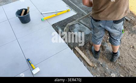 Male construction worker laying tiles on outdoor surface, using a trowel to spread adhesive, with tools and materials visible on the ground Stock Photo