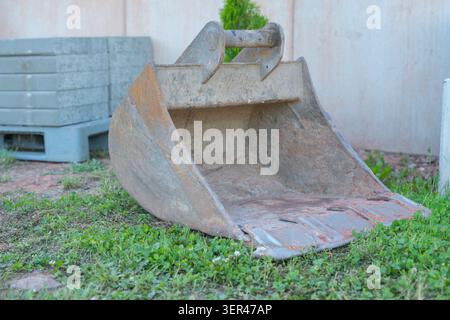 A detached rusty steel excavator bucket rests on green grass at a residential landscaping construction site during a work break. Heavy duty digging Stock Photo