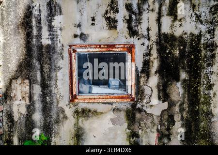 Small rusted window set in a heavily weathered concrete wall with peeling plaster, stains, and textured surfaces. Detailed architectural decay and urb Stock Photo