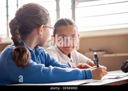 Two primary school girls sitting at their desks during class, interacting while working on school assignments. Stock Photo