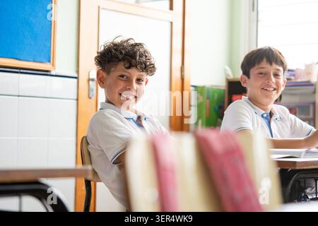 Two primary school boys smiling while sitting at their desks in a classroom. Stock Photo