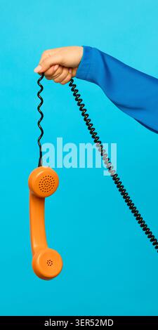 Hand holding orange telephone receiver with black cord on blue background Stock Photo